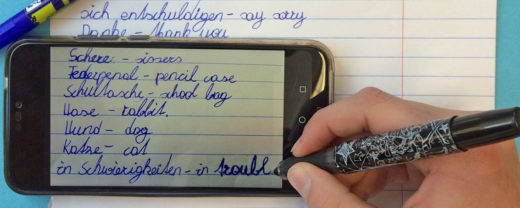 Zu sehen ist eine Hand mit einem Stift sowie ein Handy und ein Heft. Die Hand schreibt Vokabel auf englisch und deutsch. 