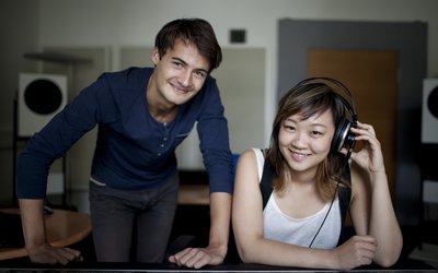 Ein Student und eine Studentin an einem Mischpult mt Kopfhörern lachen in die Kamera