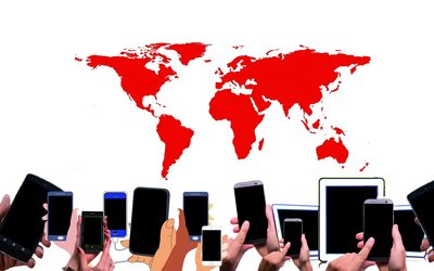 Fourteen smartphones and tablets are being held up by hands in front of a red-coloured world map in the back.