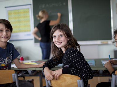 Drei sitzende Volksschulkinder in Klassenraum drehen sich von Tafel weg und blicken in die Kamera