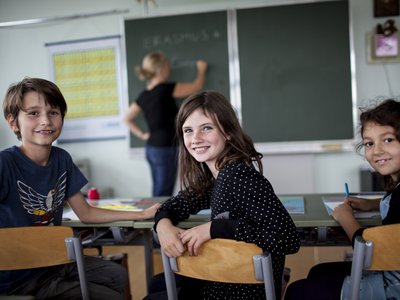 Drei sitzende Volksschulkinder in Klassenraum drehen sich von Tafel weg und blicken in die Kamera