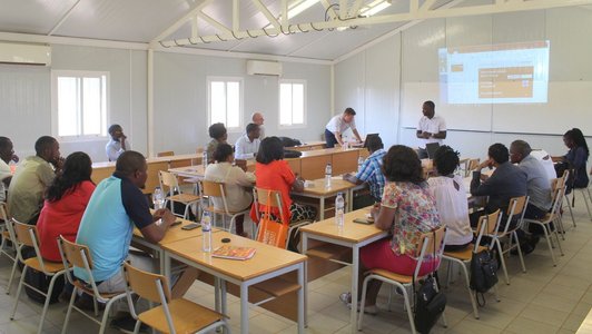 Group of people sitting in a room at tables arranged in rows and watching a presentation 