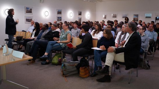 A man is giving a speech in front of an audience of approximately 50 people.