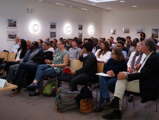A man is giving a speech in front of an audience of approximately 50 people.