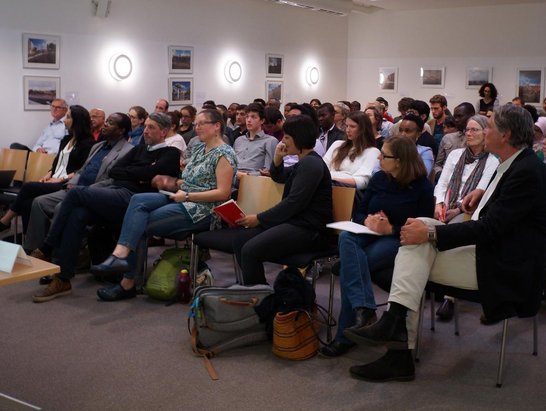 A man is giving a speech in front of an audience of approximately 50 people.