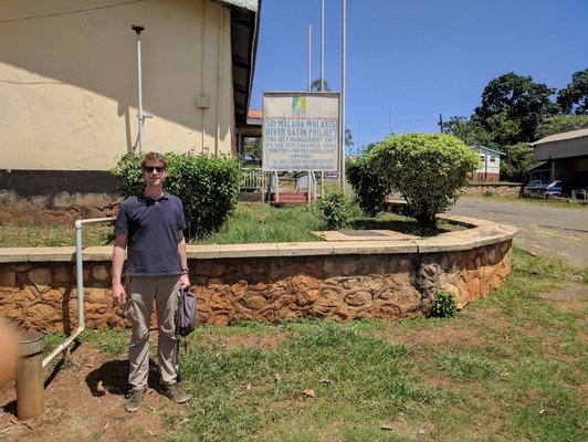 Man standing on a grassy soil in front of a beige building and a brown stone wall