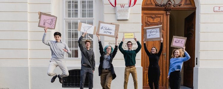 Junge Studierende hüpfen in die Luft und halten Schilder in der Hand