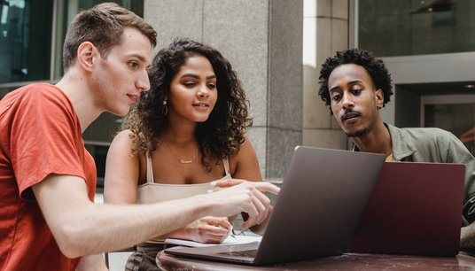 Drei Studierende sitzen am Tisch blicken auf einen Laptop