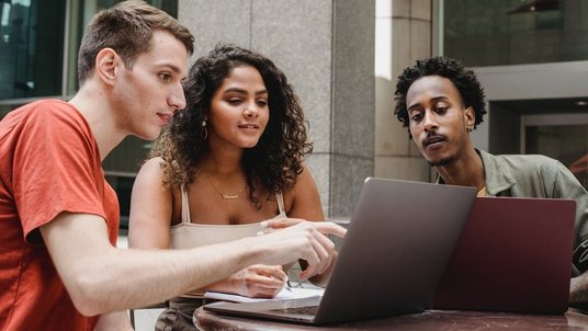 Drei Studierende sitzen am Tisch blicken auf einen Laptop