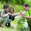Ein Forscher und zwei Mädchen hocken an einem Gewässer und beobachten Wassertiere in einem Glas.