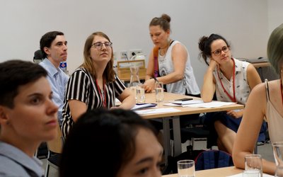 Junge Menschen blicken sitzen an Tischen in einem Seminarraum und blicken aufmerksam auf einen Punkt außerhalb des Bildraums