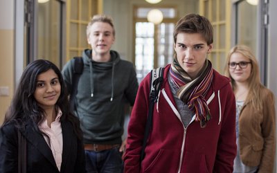 Vier Schüler und Schülerinnen gehen in einem Schulgang der Kamera entgegen