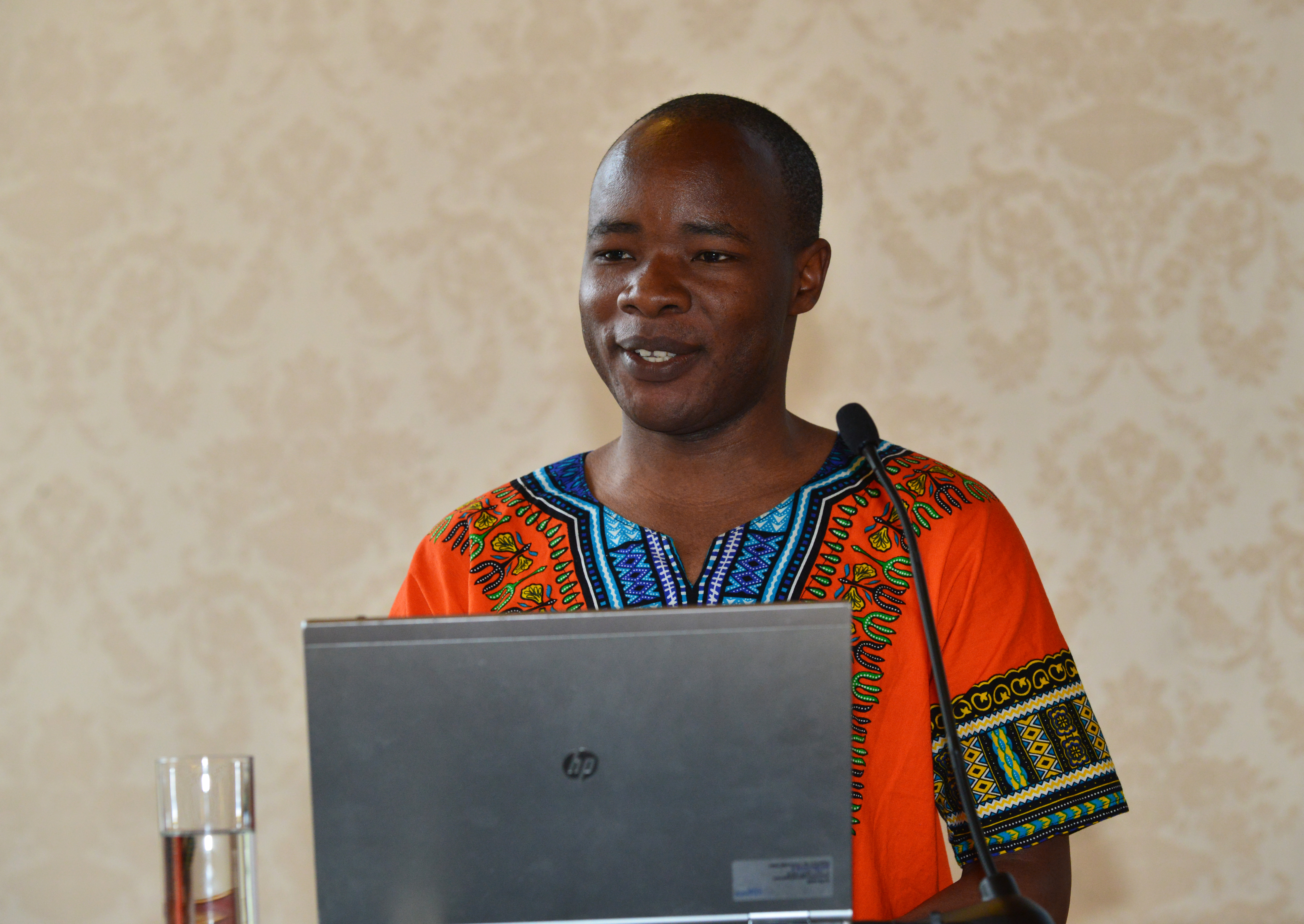 A man standing in front of a laptop, a microphone and a glass of water, apparently speaking to an audience